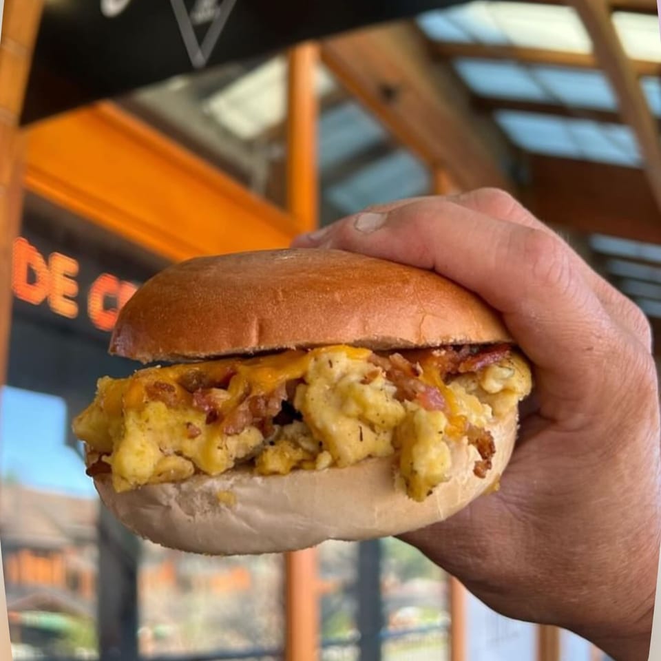 A hand holds a breakfast bagel sandwich filled with scrambled eggs, melted cheese, and bacon.