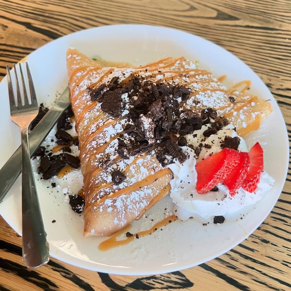 A folded crepe drizzled with caramel, topped with crushed cookies, whipped cream, and strawberries.